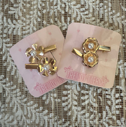 Two gold-toned hair clips with pearl details, displayed on pink 'Trendy Merch' backing cards over a textured beige lace fabric background.