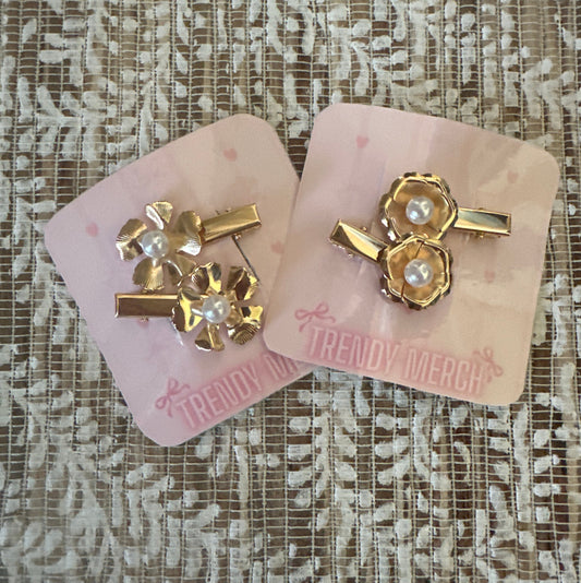 Two gold-toned hair clips with pearl details, displayed on pink 'Trendy Merch' backing cards over a textured beige lace fabric background.