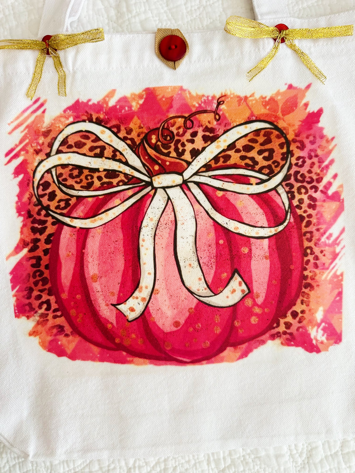 A wwhite canvas tote bag featuring a printed autumn design of a red pumpkin with a leopard-print bow and a red watercolor background, resting on a pink patterned surface.