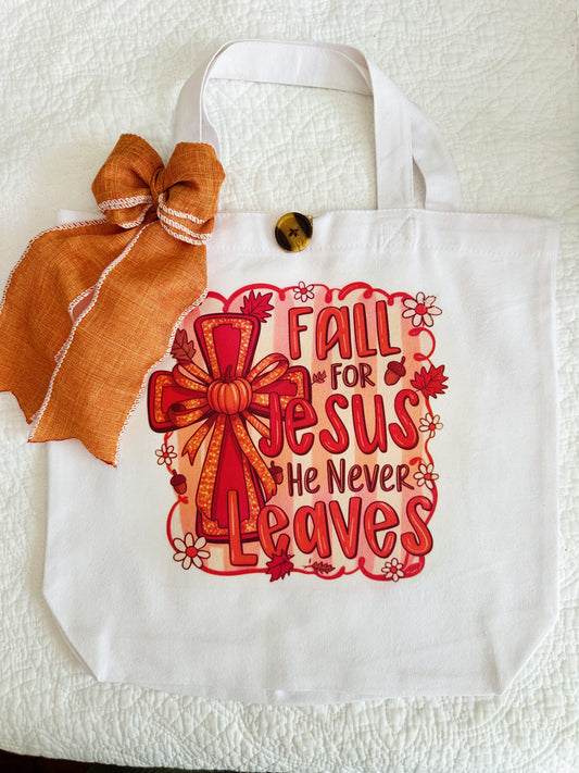 A white canvas tote bag with a decorative orange and red graphic that reads 'Fall for Jesus He Never Leaves' with a bow illustration, plus an orange ribbon bow and wooden button attached near the handle.