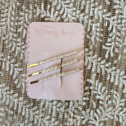 Three gold hair pins decorated with white pearls, displayed on a pink card labeled 'Fancy Nerd' and resting on a textured beige lace fabric background.