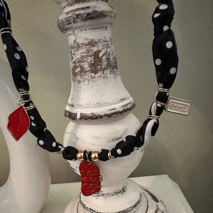 A close-up of a necklace displayed on a distressed white decorative stand. The necklace features black fabric with white polka dots, black beads, gold-toned spacer beads, and two red enamel lip-shaped charms.