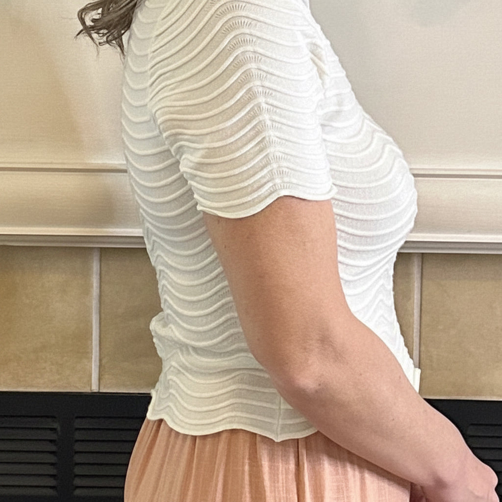Person wearing a white textured shirt and peach skirt standing against a beige tiled wall.