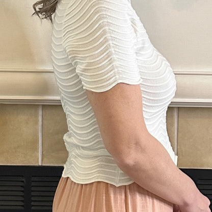 Person wearing a white textured shirt and peach skirt standing against a beige tiled wall.