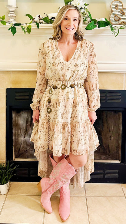 A woman stands in front of a fireplace wearing a long-sleeve, tiered cream floral dress with a decorative belt and pink cowboy boots.