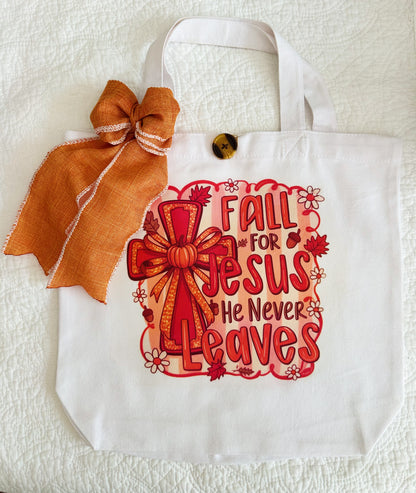 A white canvas tote bag with a decorative orange and red graphic that reads 'Fall for Jesus He Never Leaves' with a bow illustration, plus an orange ribbon bow and wooden button attached near the handle.