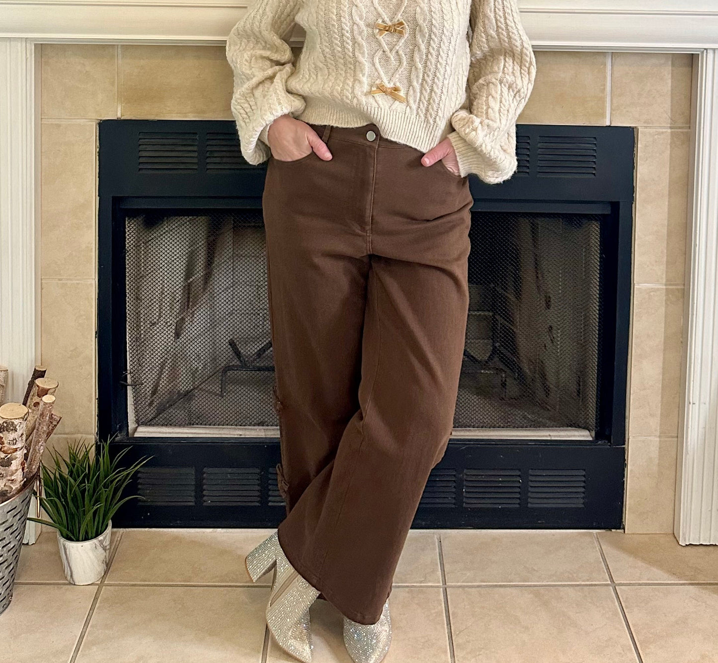 Woman standing in front of a fireplace wearing a cream sweater and brown pants.