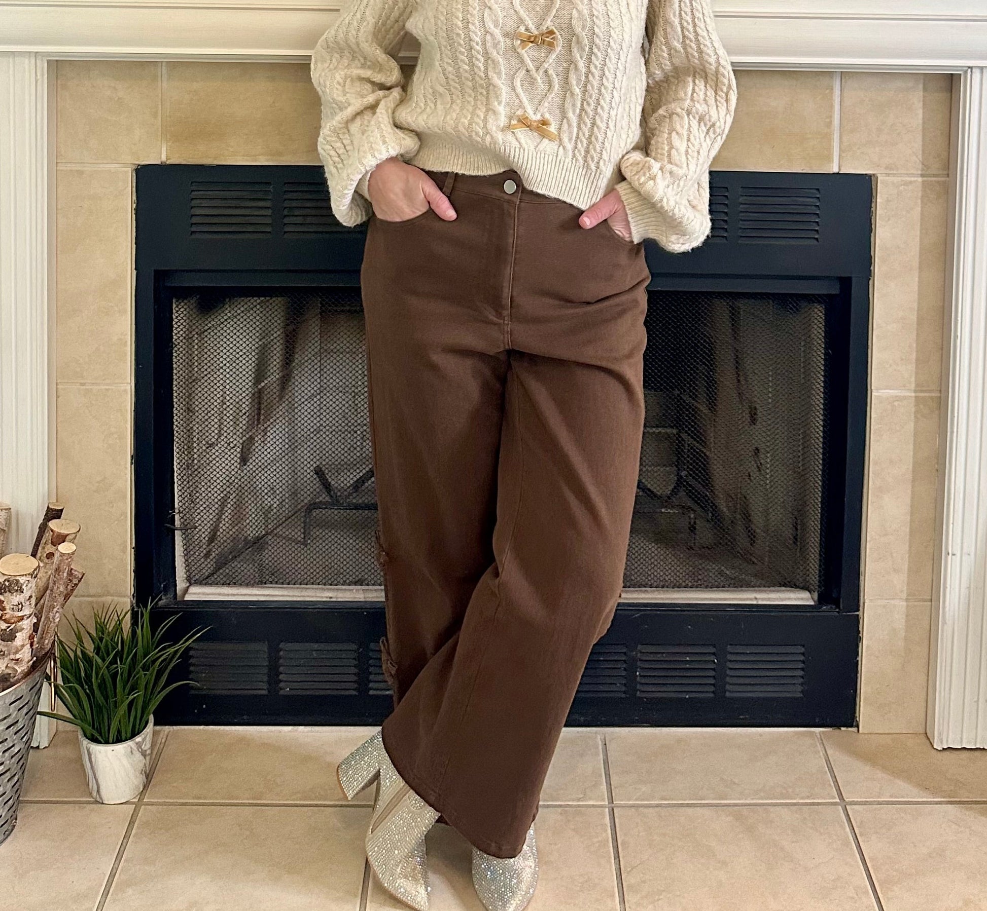 Woman standing in front of a fireplace wearing a cream sweater and brown pants.