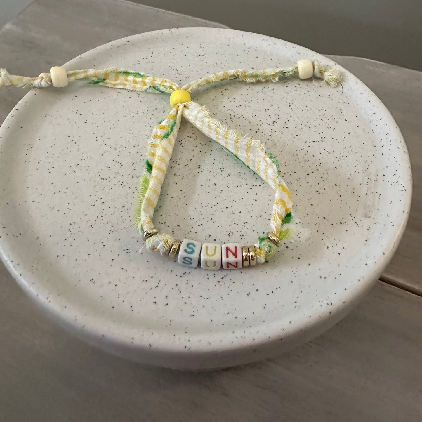 A handmade braided cord bracelet with green and yellow patterned details, a small yellow bead, and colorful letter beads spelling 'SUNNY', displayed on a speckled ceramic dish.