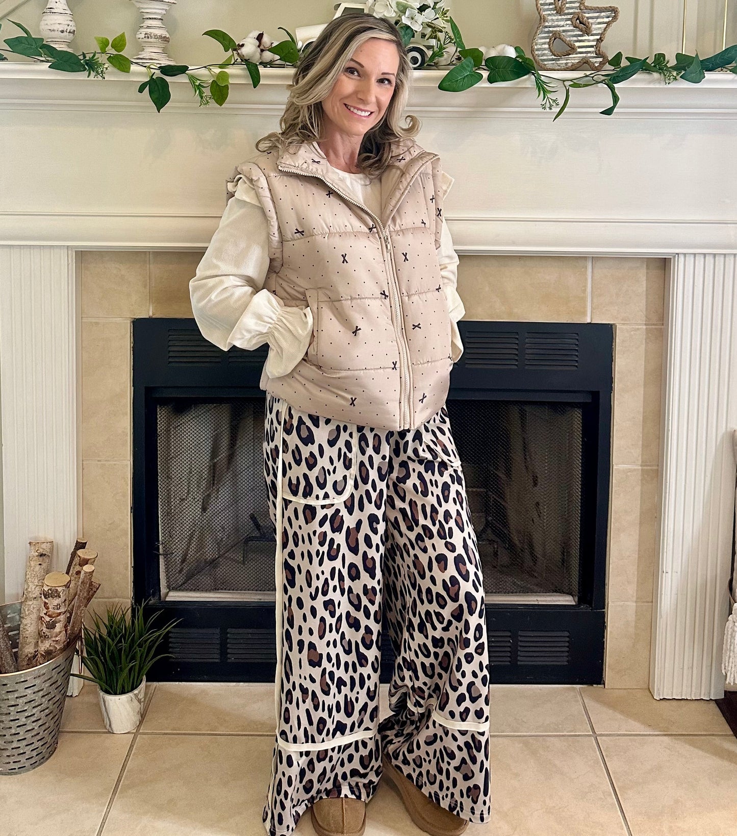 Woman in a cozy living room with a fireplace, wearing a beige vest and leopard print pants.