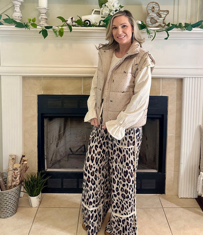 Woman in leopard print pants and beige jacket standing in a living room with a fireplace.