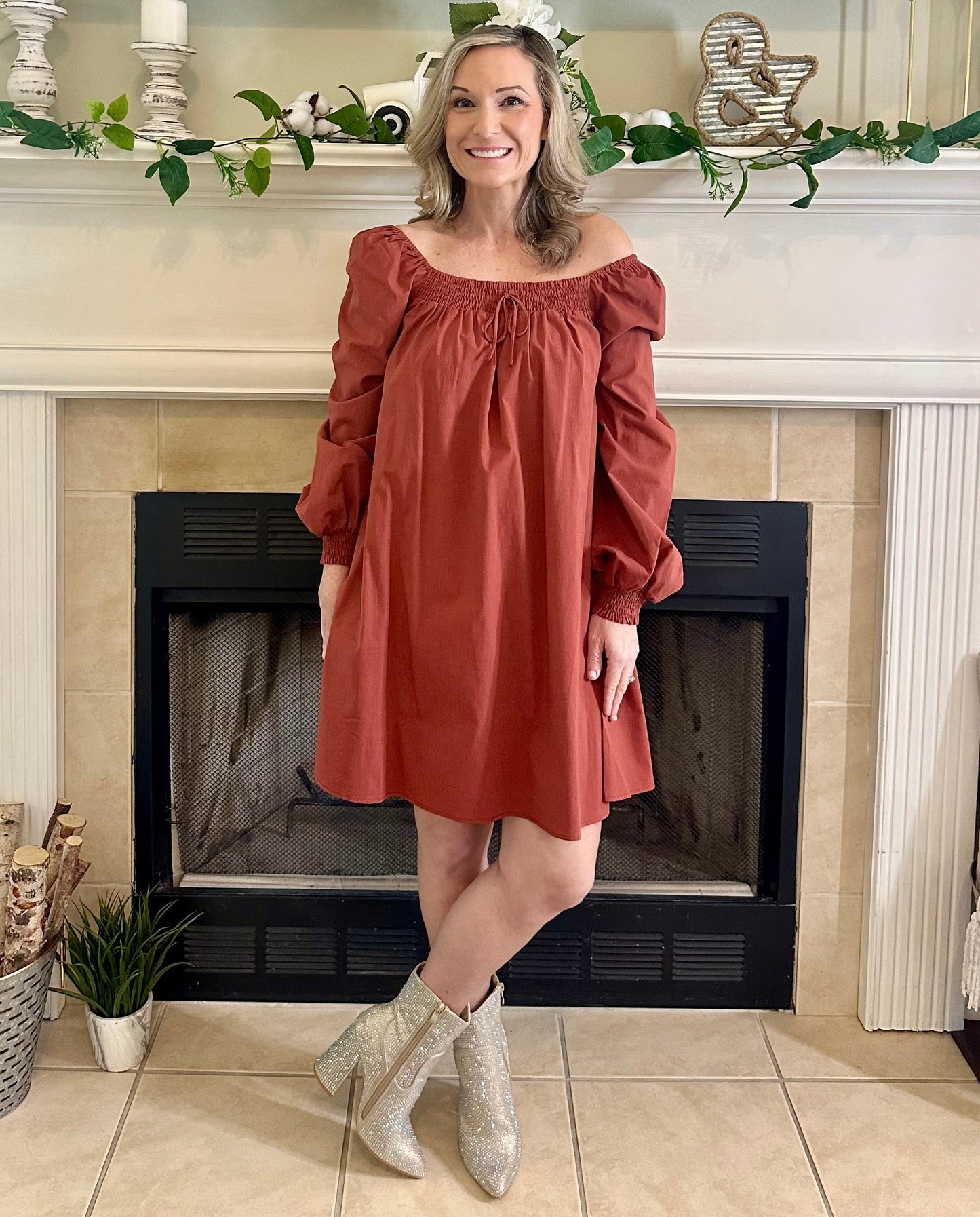 Woman in a rust-colored dress standing in front of a fireplace with decorative elements.