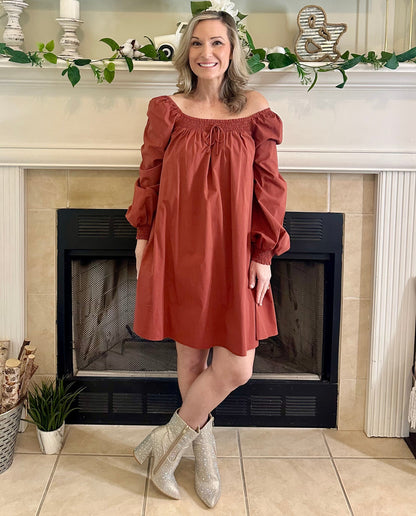 Woman in a rust-colored dress standing in front of a fireplace with decorative elements.