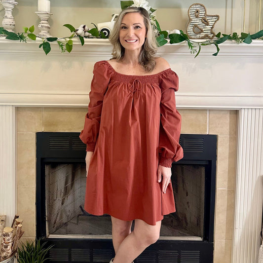 Woman in a rust-colored dress standing in front of a fireplace with decorative elements.