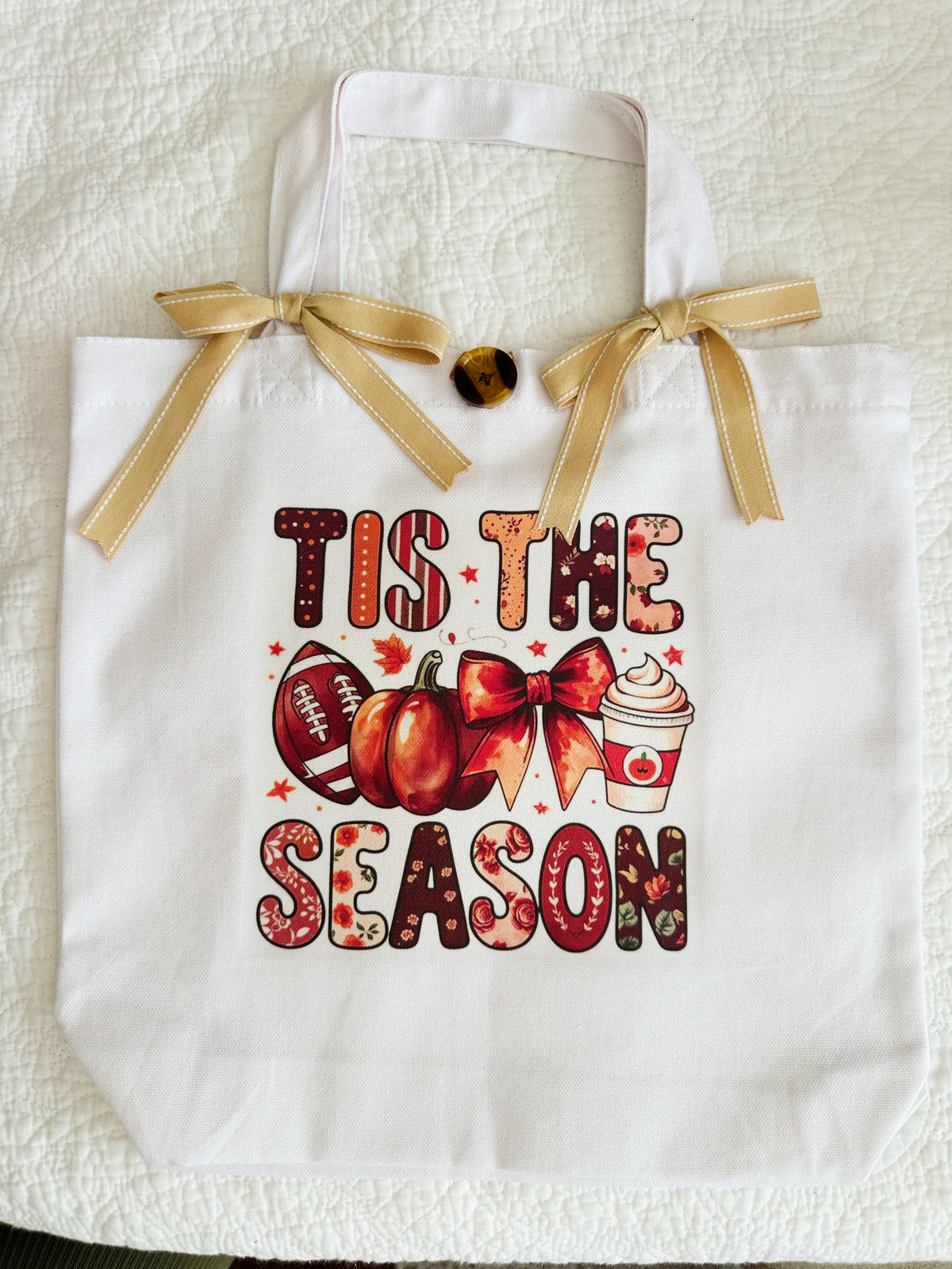 A white canvas tote bag with a gold ribbon handle bow, featuring a fall-themed graphic that reads 'TIS THE SEASON' in autumn patterned letters, alongside pumpkins, a football, a bow, and a hot cocoa cup.