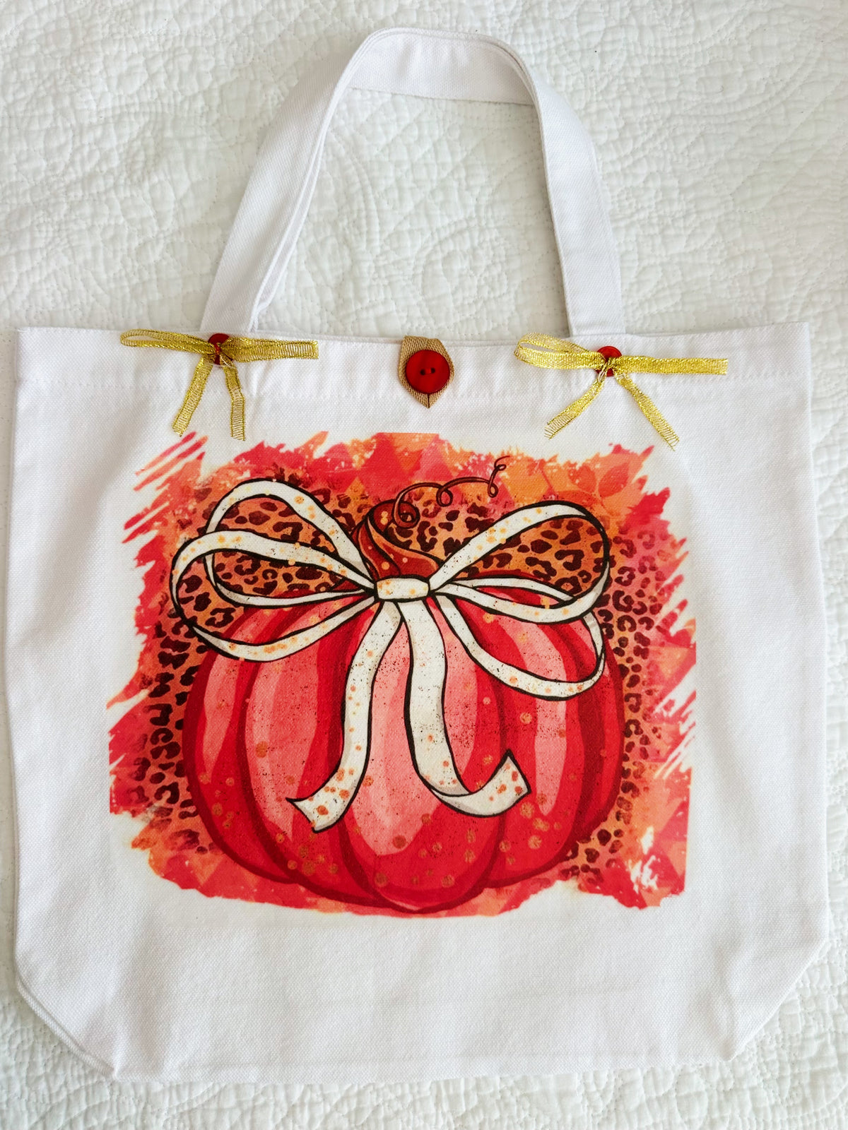A wwhite canvas tote bag featuring a printed autumn design of a red pumpkin with a leopard-print bow and a red watercolor background, resting on a pink patterned surface.