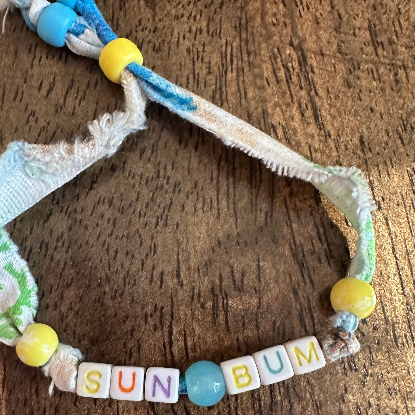 A handmade woven rope bracelet with colorful beads spelling out 'SUN BUM' on a rustic wooden surface.
