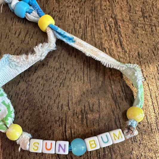A handmade woven rope bracelet with colorful beads spelling out 'SUN BUM' on a rustic wooden surface.