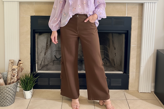 Woman standing in front of a fireplace wearing a cream sweater and brown pants.