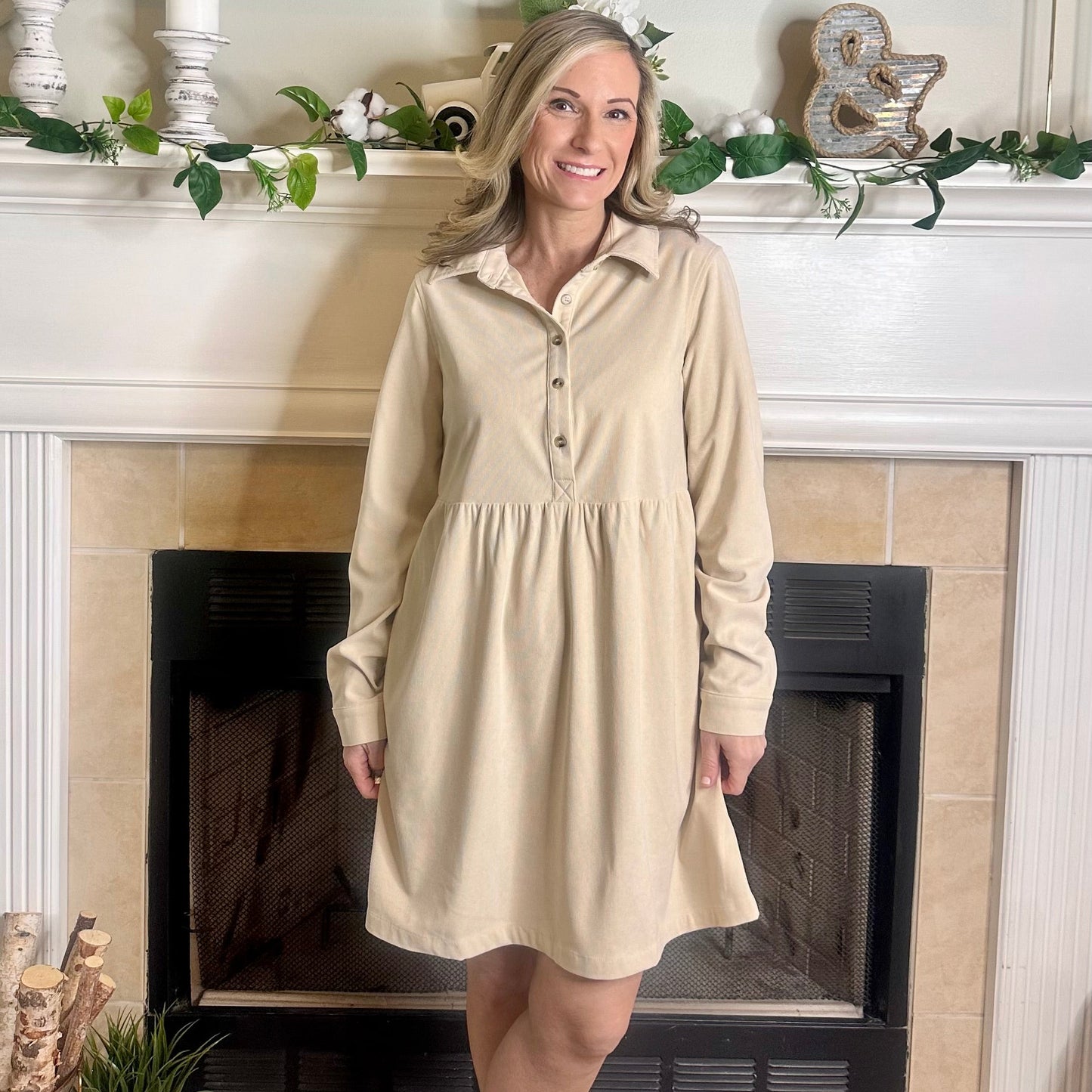 Woman wearing a beige dress standing in front of a fireplace.