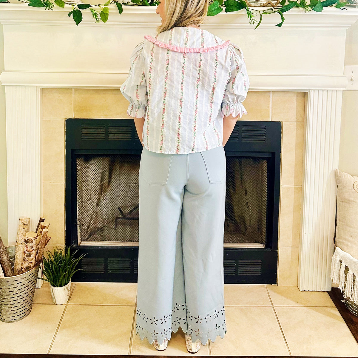 A woman stands with her back to the camera in front of a fireplace, wearing a white puff-sleeve blouse with vertical pink and green stripes and light blue wide-leg pants with eyelet detailing at the hems.
