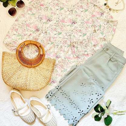 A styled flat-lay outfit featuring a cream floral print top, sage green laser-cut pants, white Mary Jane shoes, a woven straw handbag, and sunglasses on a textured white blanket.