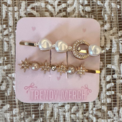 A set of  gold-toned hair clips decorated with faux pearls and crystals, attached to a pink display card labeled 'TRENDY MERCH' and placed on a textured beige lace fabric background.