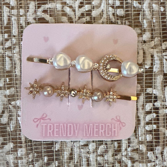 A set of  gold-toned hair clips decorated with faux pearls and crystals, attached to a pink display card labeled 'TRENDY MERCH' and placed on a textured beige lace fabric background.