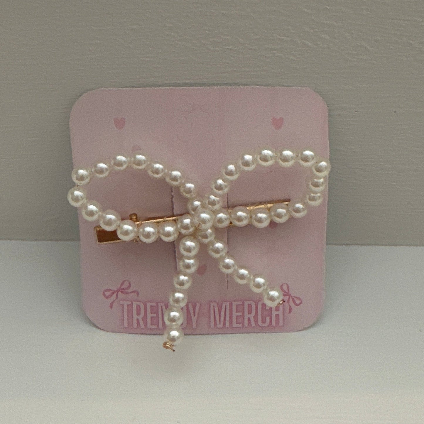 A pearl-bead bow hair clip with a  gold metal clip, displayed on its pink backing card which reads 'TREZY MERCH'.