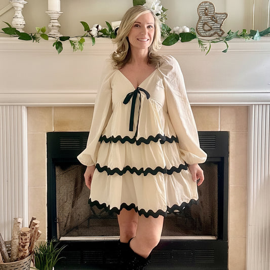 Woman in a cream dress with black trim standing in front of a fireplace.