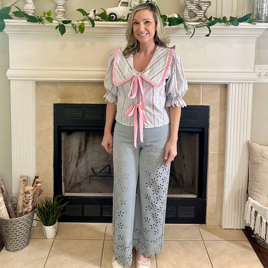 Woman standing in a living room with a fireplace, wearing a patterned top and light blue pants.