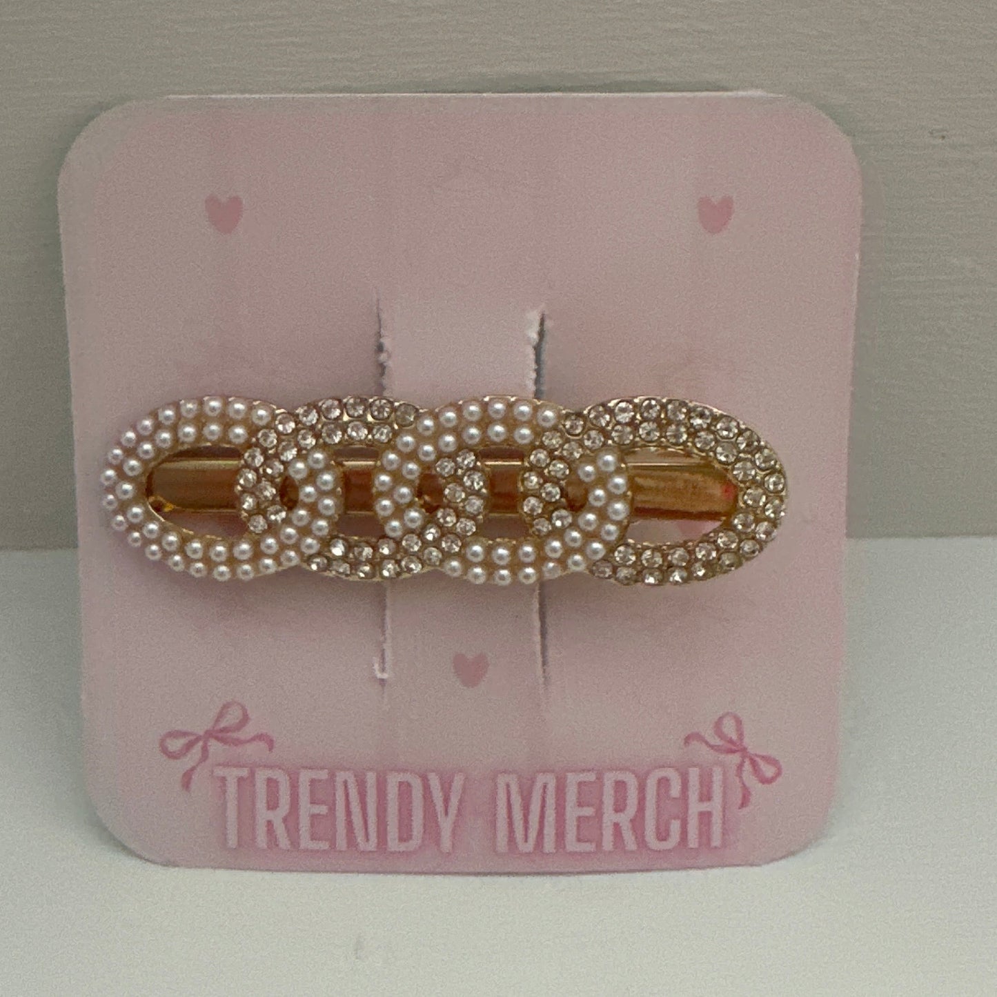 A gold-tone hair claw clip embellished with white faux pearls, displayed on a pink card labeled 'TRENDY MERCH' against a neutral gray background.
