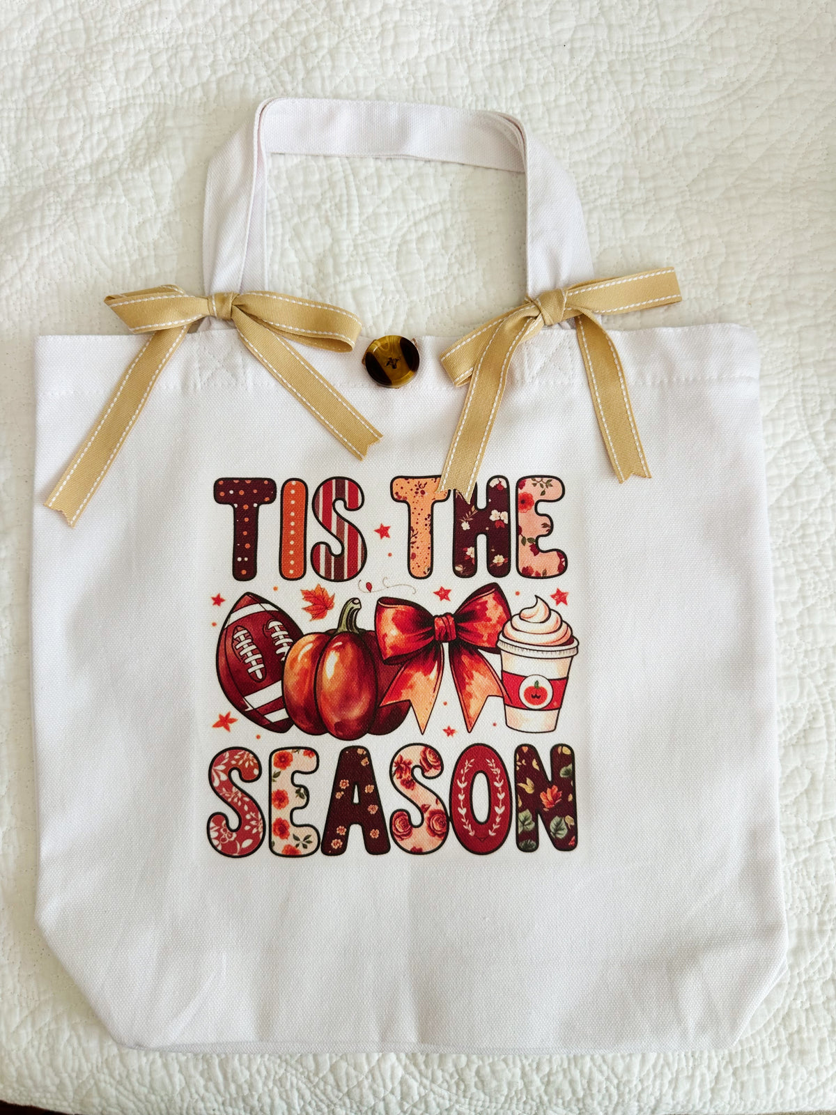 A white canvas tote bag with a gold ribbon handle bow, featuring a fall-themed graphic that reads 'TIS THE SEASON' in autumn patterned letters, alongside pumpkins, a football, a bow, and a hot cocoa cup.