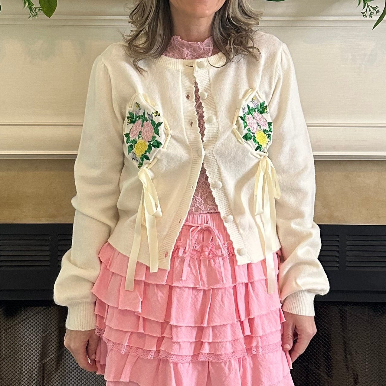 Person wearing a cream cardigan with floral embroidery and a pink skirt in front of a fireplace.