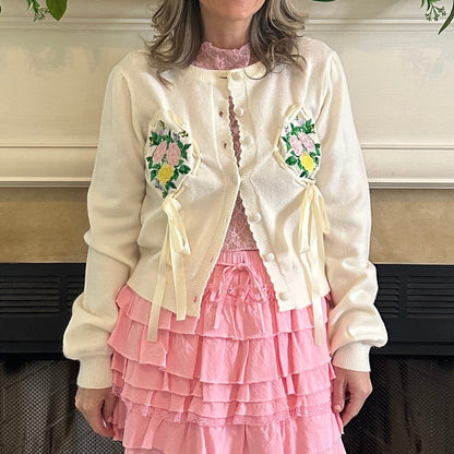 Person wearing a cream cardigan with floral embroidery and a pink skirt in front of a fireplace.