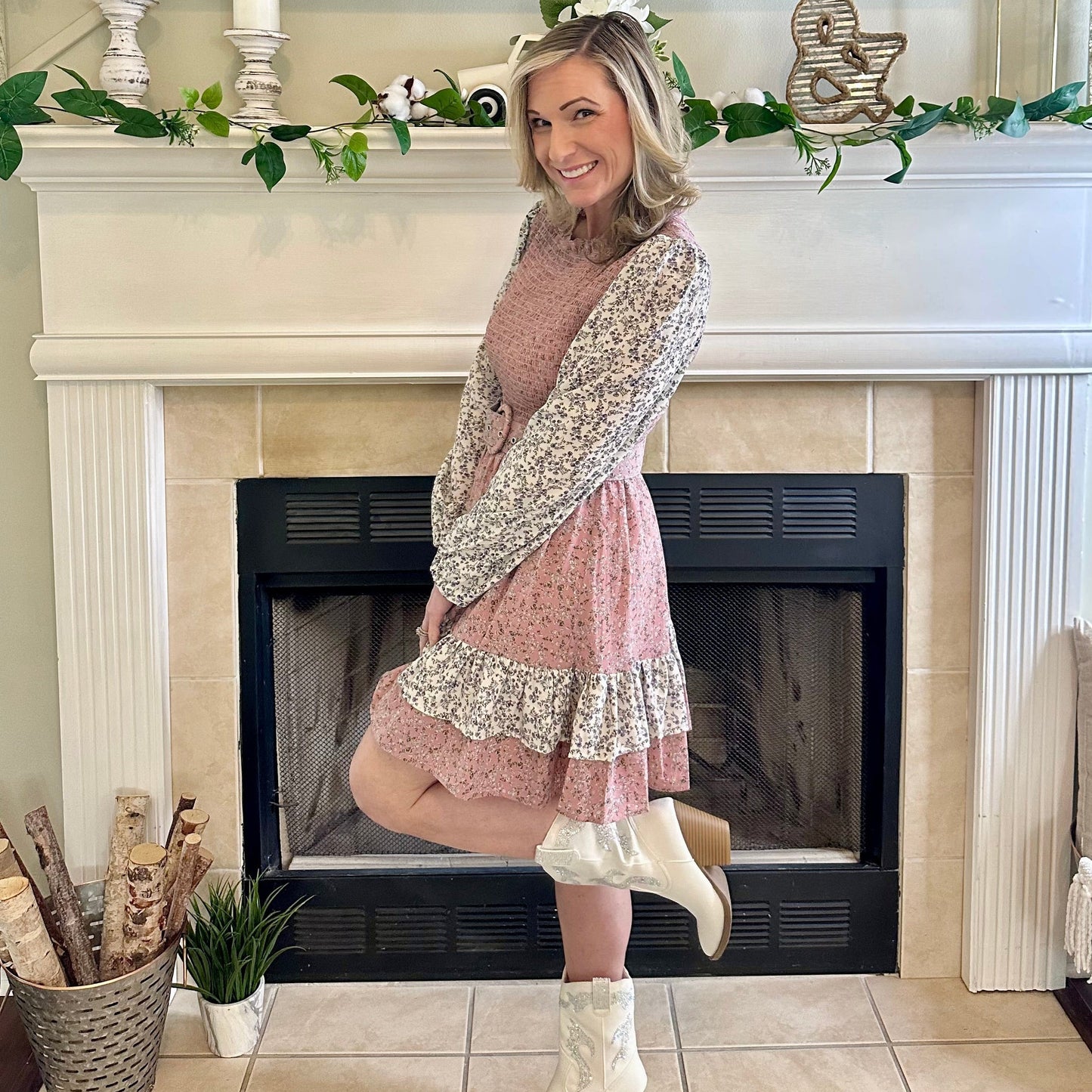 Woman in a floral dress and white boots standing in front of a fireplace.