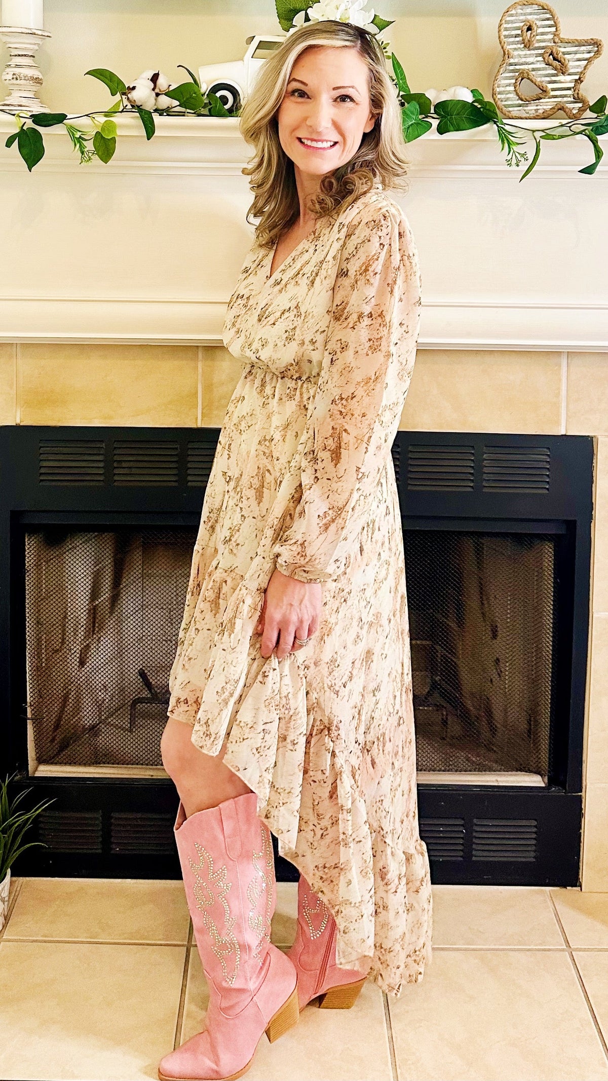 A woman stands in front of a fireplace wearing a long-sleeve, high-low floral dress in a light beige and pink tone, paired with pink knee-high boots.