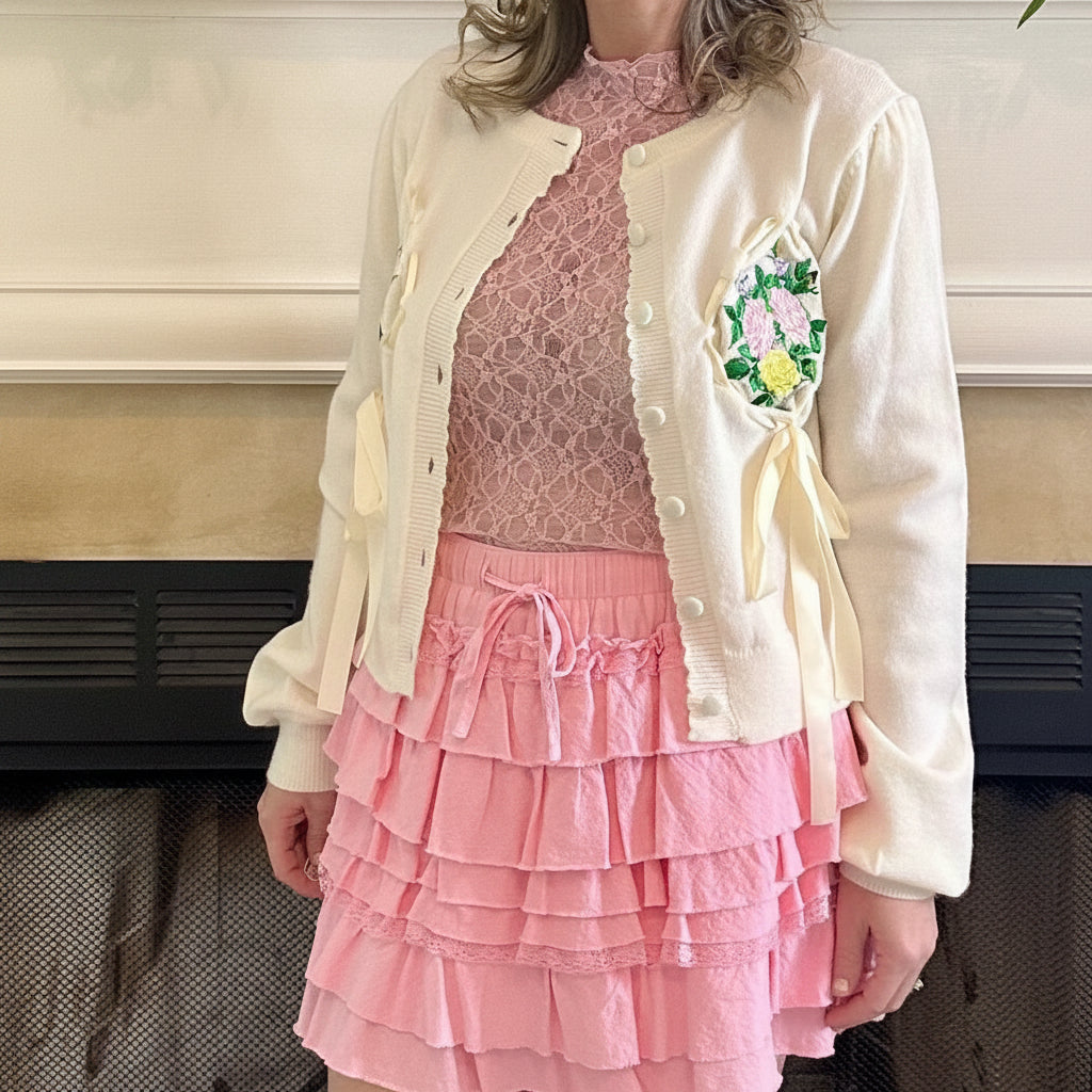 Woman in a pink dress with a white jacket standing in front of a fireplace.