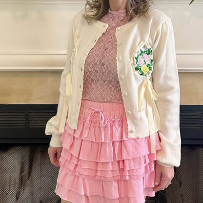 Woman in a pink dress with a white jacket standing in front of a fireplace.