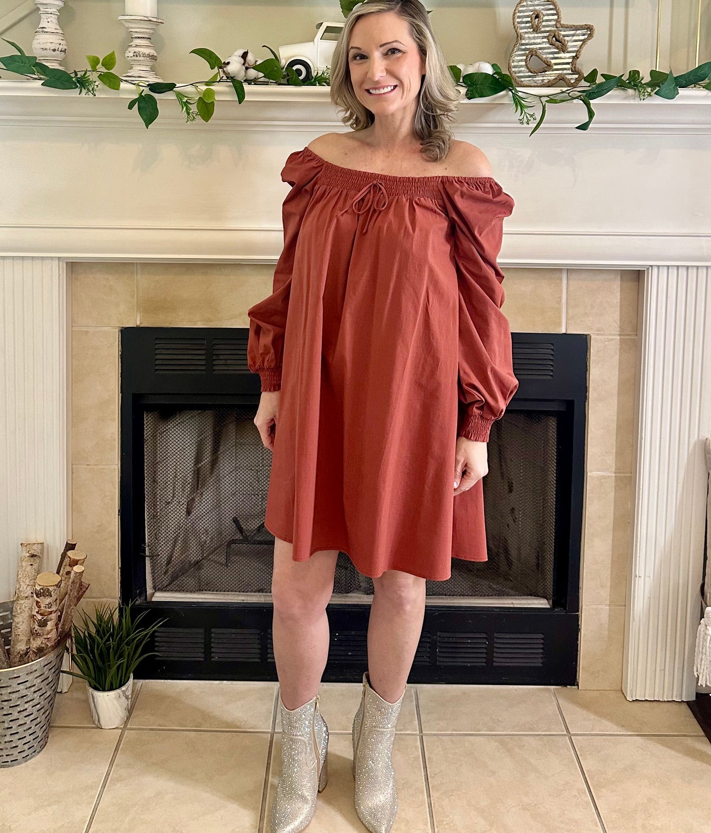 Woman in a rust-colored off-shoulder dress standing in front of a fireplace.