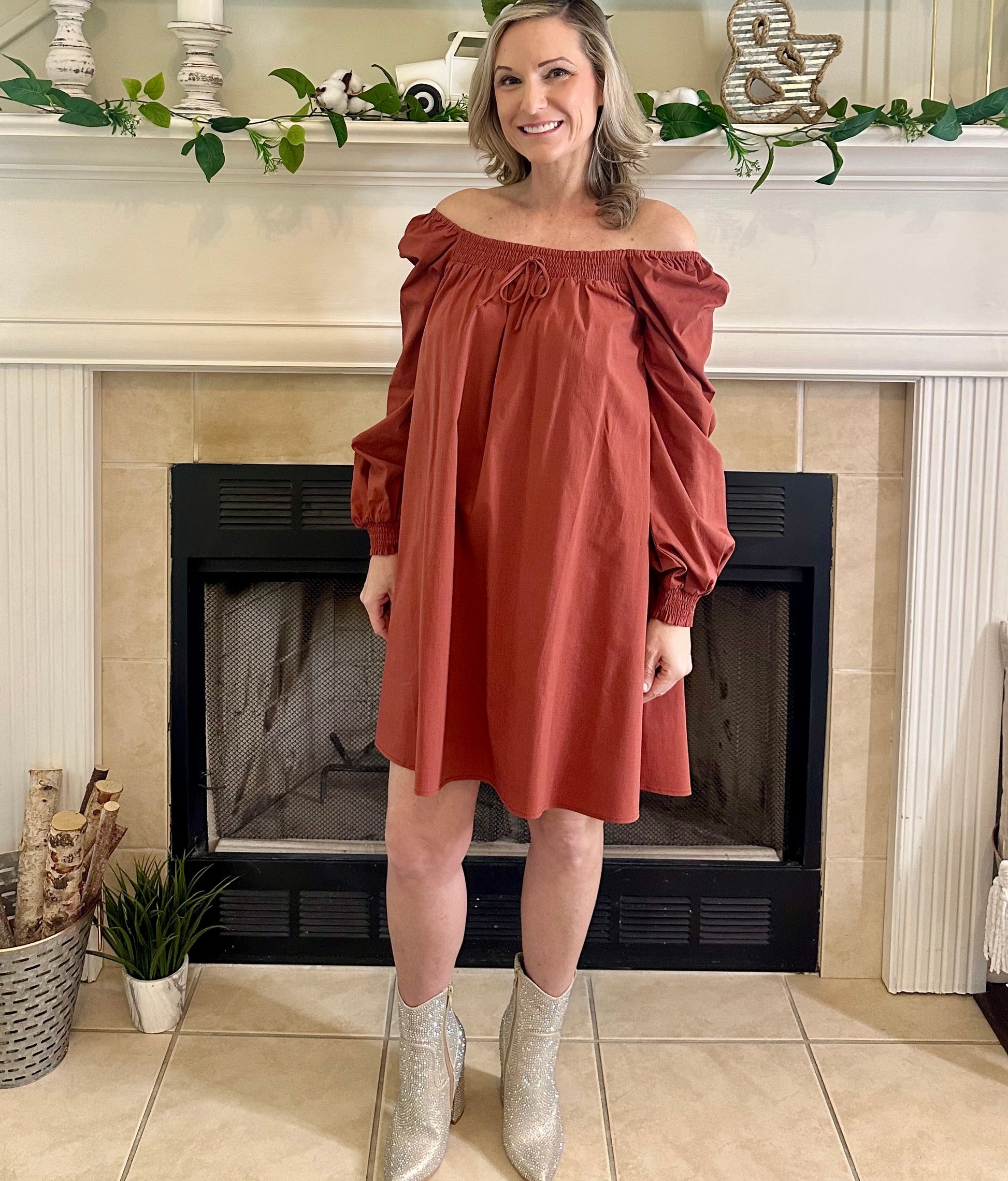 Woman in a rust-colored off-shoulder dress standing in front of a fireplace.