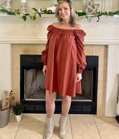 Woman in a rust-colored off-shoulder dress standing in front of a fireplace.