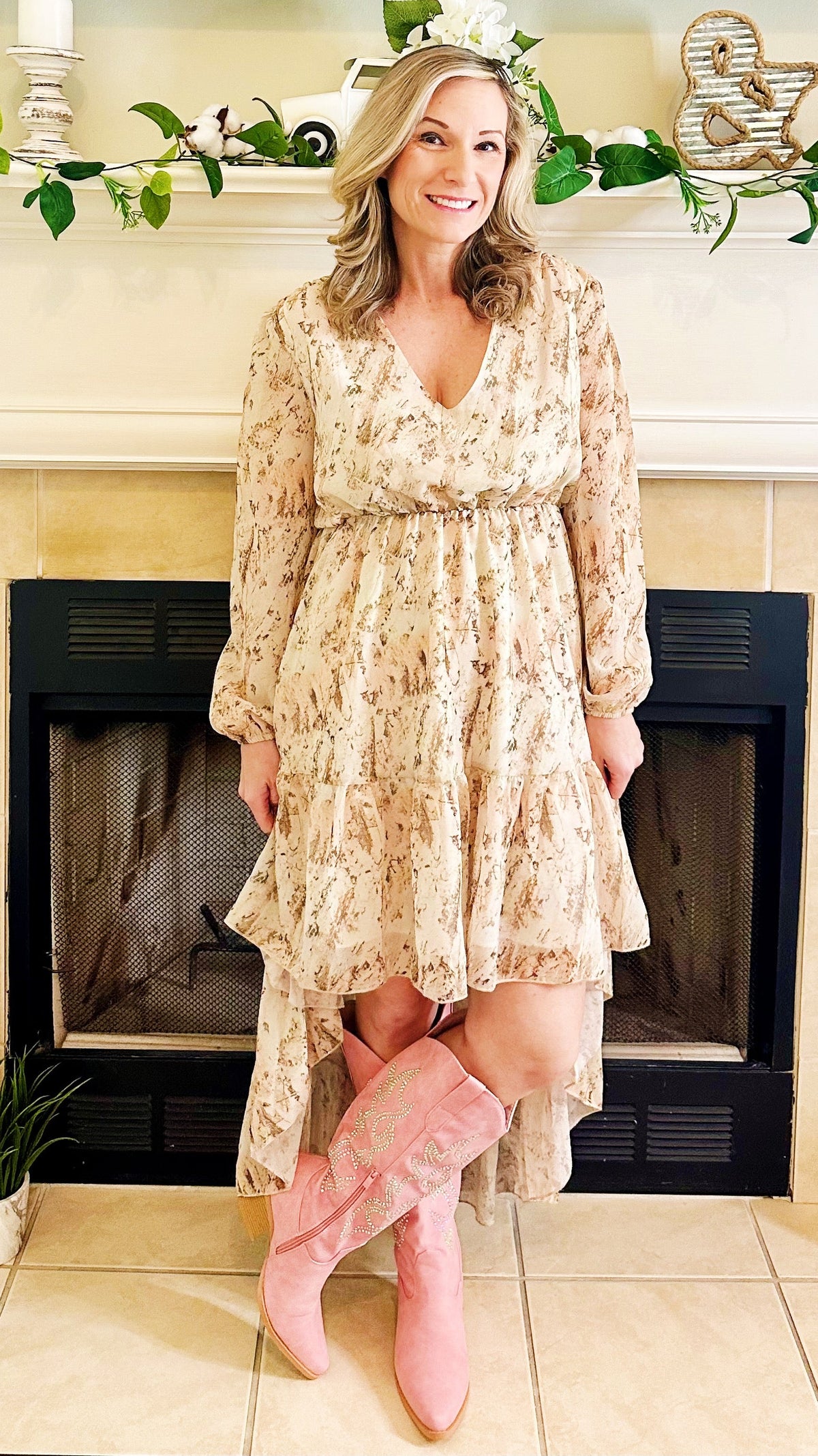 A woman stands in front of a fireplace wearing a long-sleeve, high-low floral dress in a light beige and pink tone, paired with pink boots.