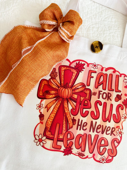 A white canvas tote bag with a decorative orange and red graphic that reads 'Fall for Jesus He Never Leaves' with a bow illustration, plus an orange ribbon bow and wooden button attached near the handle.