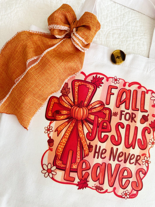 A white canvas tote bag with a decorative orange and red graphic that reads 'Fall for Jesus He Never Leaves' with a bow illustration, plus an orange ribbon bow and wooden button attached near the handle.