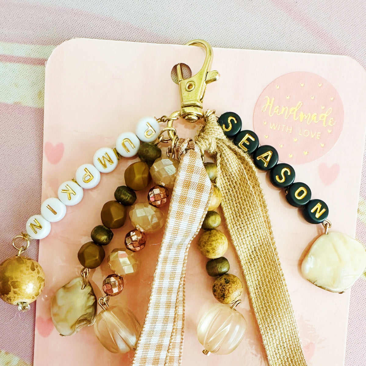A wbohemian-style beaded keychain charm with wooden beads, clear glass beads, a gold clasp, a 'SEASON' bead string, and two hanging ribbons, displayed on a dark wood background.