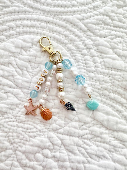 A beaded keychain with a gold lobster clasp, featuring white faux pearls, light blue beads, a starfish charm, a seashell charm, and a striped shell charm, displayed on a white quilted blanket.