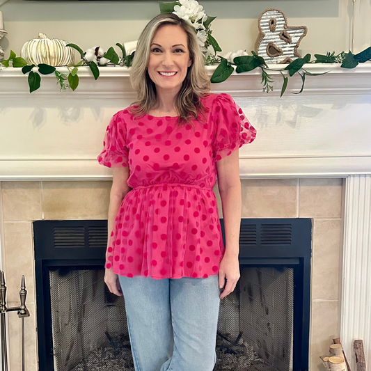 Woman wearing a pink polka dot blouse and blue jeans standing in front of a fireplace.