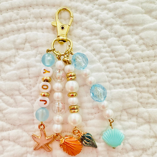 A gold-tone lobster clasp keychain bag charm featuring white faux pearls, light blue beads, a 'JOY' bead section, and sea-themed charms including a starfish, orange seashell, and turquoise seashell, displayed on a textured white fabric background.