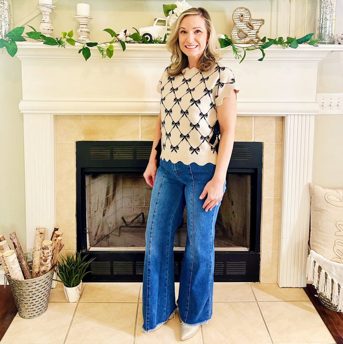 A woman stands in front of a fireplace wearing a cream short-sleeve top with a black bow pattern and blue wide-leg jeans.